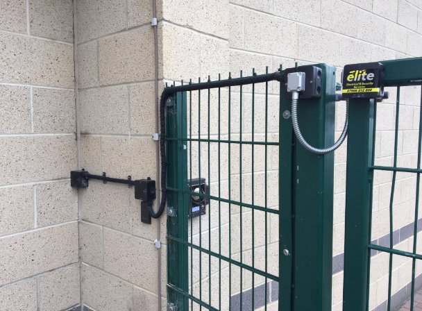 A magnetic lock fitted to a green mesh security gate, with its cable run up the wall